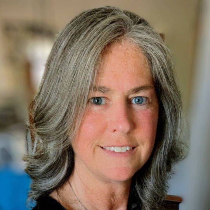 A woman with short gray hair and a floral print black tank top smiling in front of an out-of-focus indoor background